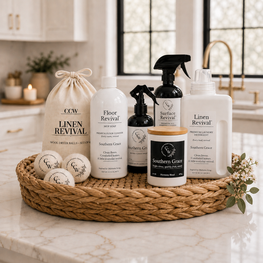 CCW  Home Revival Set featuring Southern Grace scented laundry detergent, mop soap, room and linen spray, Surface Revival all-purpose cleaner, wool dryer balls, and candle displayed on a woven tray in a bright farmhouse kitchen.